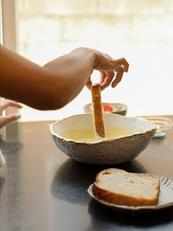 A person dips a slice of bread into a large bowl of soup or vegan French toast batter, with more bread on a plate nearby on a kitchen counter. Sunlight streams in from a window in the background.